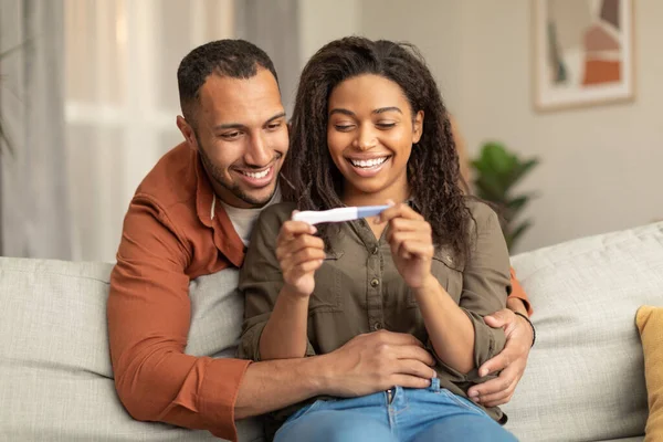 幸せなアフリカ系アメリカ人のカップルの肯定的な妊娠テストと笑顔を保持し 自宅でソファの上に男の巨大な女性 黒人の配偶者は赤ちゃんについて喜んで学ぶ — ストック写真