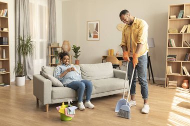Pregnancy and household help. Black husband cleaning home while his pregnant wife resting and sitting on sofa at home. Family lifestyle and housework leisure concept