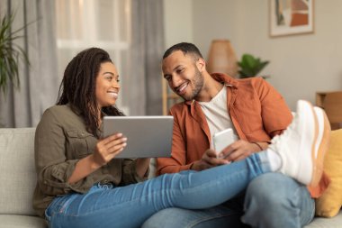 Relaxed black spouses chilling together at home, happy man and woman using digital tablet and smartphone, resting on sofa. Entertainment and gadgets concept