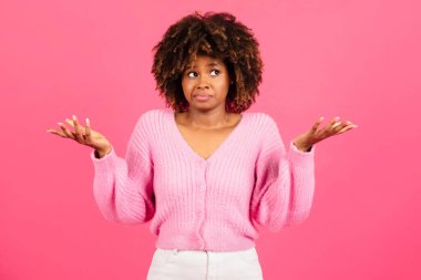 Insecure confused incompetent millennial african american curly lady in casual spreads her arms to sides and holds empty space isolated on pink background, studio. Human emotions, choice, question