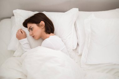 Upset young brunette woman lying alone with open eyes in big bed at home, unwilling to wake up or cant sleep, depressed lady suffering from insomnia, top view, copy space for ad
