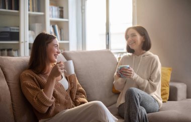 Happy european young women in casual with cups drink coffee, communication, sit on sofa in living room interior. Visit to friend, meeting together, bachelorette party with gossip, friendship at home