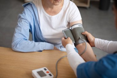 Doctor measures blood pressure with tonometer of senior european female patient in clinic office interior, cropped. Health care, treatment of high pressure, visit to therapist and medical examination