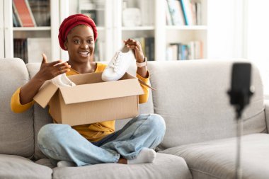 Social media influencer stylish cheerful young black woman reviewing product. Excited african american millennial lady vlogging about unpacking white sneakers and filming herself at home on cell phone