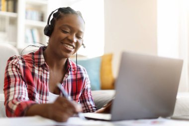 E-learning, online school, education. Joyful smart young black woman student attending webinar or online training from home, sitting on floor, using laptop and headset, taking notes, copy space
