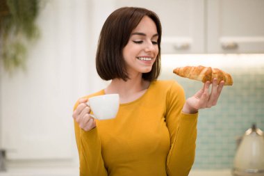 Joyful pretty young lady with nice hairstyle in casual longsleeve having breakfast at home, drinking aromatic coffee and eating croissant homemade pastry, kitchen interior, copy space