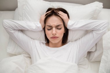 Top view of stressed sad irritated young brunette woman wearing white pajamas lying in bed at home, touching head with both hands, feeling tired after sleepless night. Insomnia, sleep disorder conept