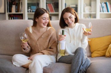 Laughing european young girlfriends with glasses and bottle of wine talking, enjoy spare time, sitting on sofa, have fun in living room interior. Visit to friend, bachelorette party together at home
