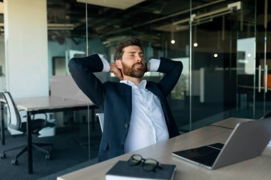 Relaxed male entrepreneur resting with closed eyes at workplace in office, leaning back in chair with hands behind head, having break after working on laptop