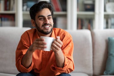 Cool handsome middle eastern millennial man in smart casual sitting on couch in living room, enjoying fresh morning coffee at home, smiling, looking at copy space. Domestic lifestyle concept