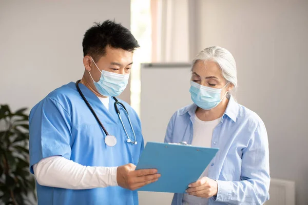 Serious middle aged korean doctor in protective mask giving contract sign to old caucasian female patient in clinic office interior. Visit to therapist and medical examination, health care, treatment