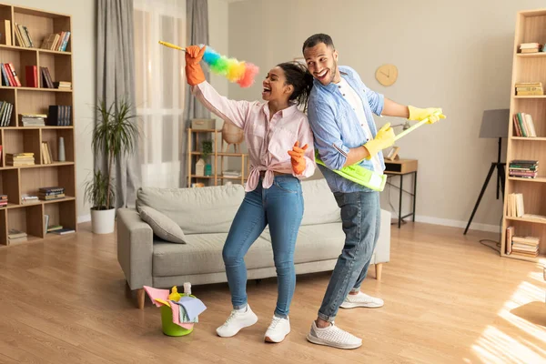 Happy joyful black husband and wife tidying home and singing during housecleaning, using mop and duster as microphones. Excited spouses enjoying domestic chores