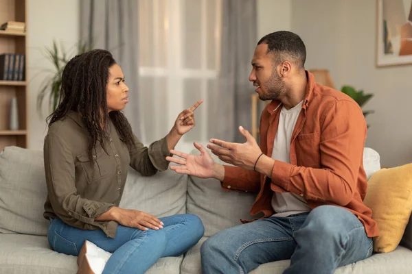 Emotional african american husband and wife sitting on couch, screaming at each other and gesturing, having fight at home. Crisis in relationships concept
