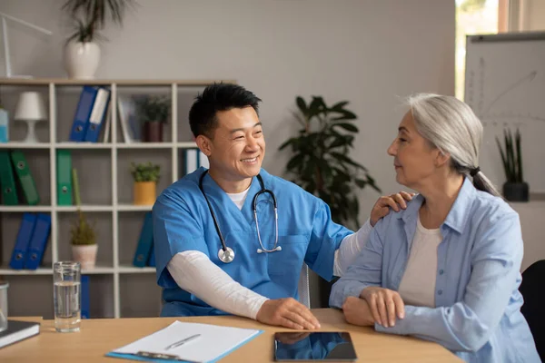 Cheerful middle aged korean man doctor supports and advises, calms old european female patient in clinic office interior. Greeting, help, health care, treatment and visit to therapist, medical