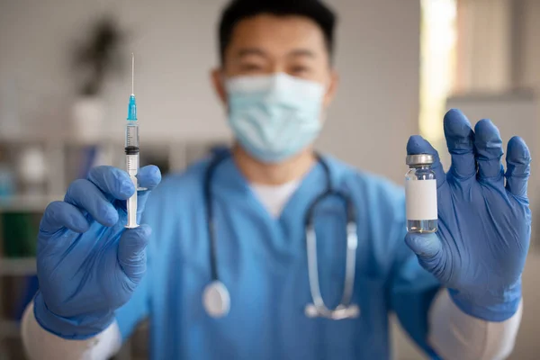 Cheerful mature korean doctor in uniform with stethoscope in protective mask shows syringe, vaccine in clinic interior. Health care, vaccination and immunization, flu and cold, due covid-19 pandemic
