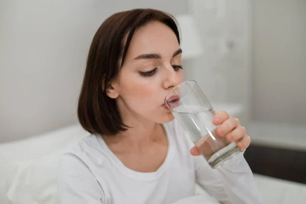 Closeup of healthy young brunette woman in white pajamas sitting in bed, drinking fresh water after waking up in the morning, copy space, side view. Hydration, healthy lifestyle concept