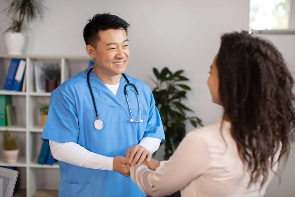 Cheerful middle aged korean doctor shaking hands, greets and supports black millennial female patient in clinic office interior. Thanks, health care, visit to family therapist and treatment of ill