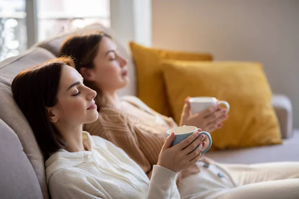 Calm european young women in casual with closed eyes relaxing, enjoy cups of tea, silence and comfort, sit on sofa in living room interior. Visit to friend, coffee break, rest and meditation at home