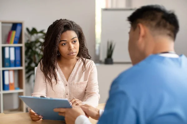 Middle aged korean doctor in uniform consults sad young african american female shows tablet in clinic office interior. Contract, health care, patient examination and medicine with family therapist