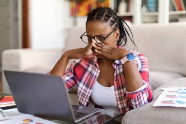 Overworked exhausted young black woman freelancer sitting on floor in front of laptop, rubbing her face, removing eyeglasses, feeling tired, home interior, copy space. Burnout in millennials concept