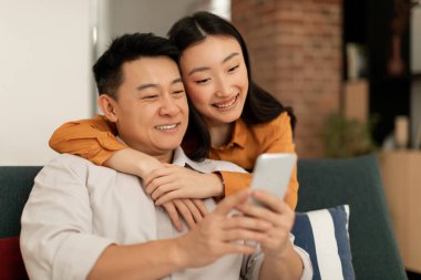Happy asian man and woman sitting on couch, using smartphone, looking at cellphone screen and smiling, using newest mobile application, home interior, copy space