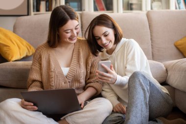 Smiling european young women with laptop and smartphone surfing in internet, chatting, show video, have fun, enjoy free time in living room interior. Visit to friend, app for social networks at home