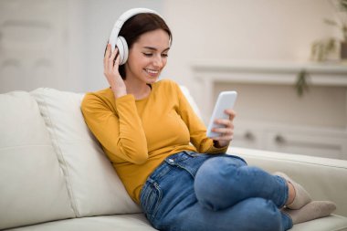 Positive smiling pretty brunette young woman in casual sitting on couch, using white wireless headphones and brand new cell phone, having online party with friends while staying home, copy space