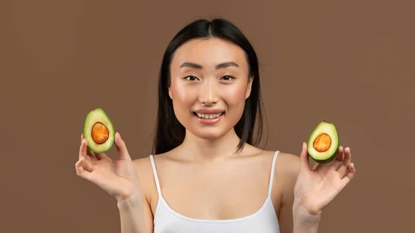 Organic skincare. Happy asian lady holding avocado halves in hands and smiling at camera, enjoying essential oils for beauty treatments, brown background, panorama