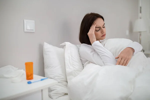 Young sick brunette woman with fever sitting in bed under warm blanket at home, touching her head, lady got coronavirus, copy space. Cold, flu, coronavirus, influenza concept