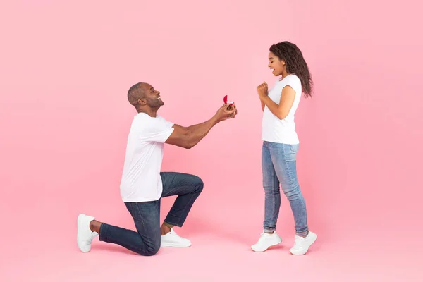 可爱的中年黑人男子单膝站立 向他心爱的年轻女子献上订婚戒指 粉红的演播室背景 侧面的景色 男人向情人求婚 — 图库照片