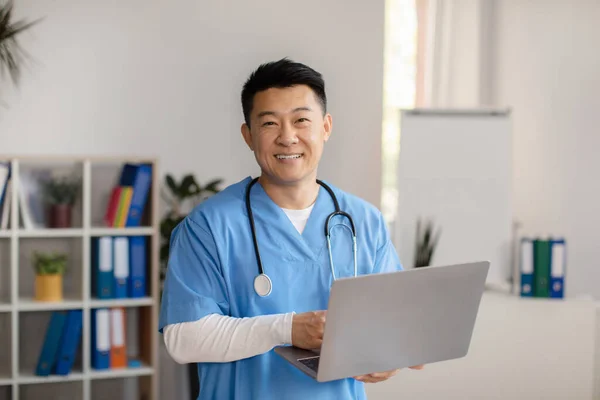 Cheerful middle aged korean doctor in uniform with stethoscope with computer in clinic office interior. Video call for medical help, remote examination, new normal and consultation, treatment disease