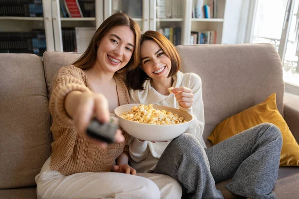 Happy european young women with remote control eat popcorn, have fun, enjoy free time, watch film in living room interior. Visit, bachelorette party, entertainment, movie evening together at home