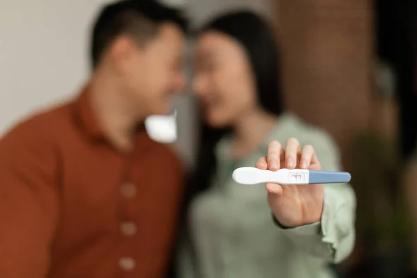 Loving asian couple with positive pregnancy test embracing, woman showing test to camera, selective focus. Korean family enjoying parenthood, hugging at home