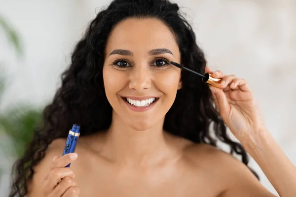 Portrait of smiling muslim millennial curly woman applying mascara on eyelashes looking in camera in bedroom interior, close up. Daily treatments, cosmetics and nude makeup beauty care and lifestyle