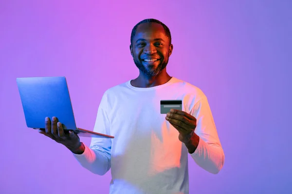 幸せなひげそりハンサムな成熟したアフリカ系アメリカ人男性でホワイト開催現代のラップトップとクレジットカードネオンスタジオの背景 カメラで笑顔 インターネット上のショッピング 購入商品 コピースペース — ストック写真