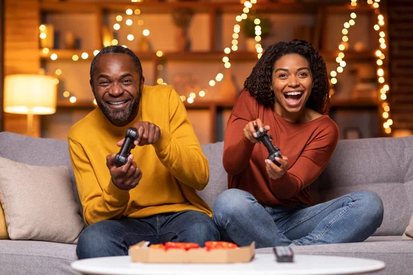Emotional happy black couple sitting on couch, holding joysticks and laughing, man and woman playing video games at home, eating pizza, living room decorated with festive lights, copy space