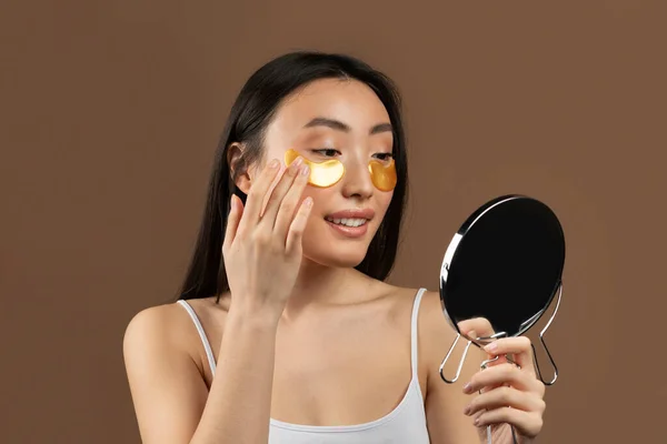 Young asian woman with natural beauty applying patches under her eyes and looking in mirror, brown background, studio shot. Beauty care, anti-aging procedures, fight against old skin