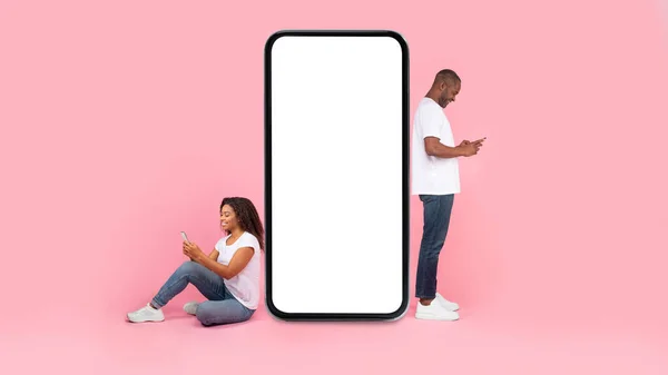 Cool application. Black couple near giant smartphone with blank white screen using gadgets, chatting or surfing social networks, pink studio background, mock up, full body length