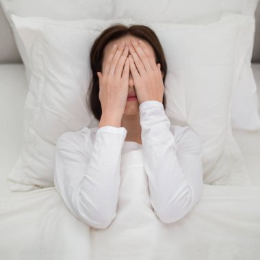 Top view of unhappy brunette young woman in white pajamas lying in white bed at home, covering her face from morning light with palms. Depression, anxiety, mental health concept