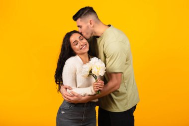 Smiling young arab male kissing, hugging woman with bouquet of flowers, congratulations with anniversary, isolated on orange background, studio. Holiday, celebration birthday, love and romance date