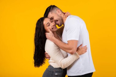 Happy attractive young middle eastern woman and man in casual hugging, kiss, enjoy love isolated on orange background, studio. Relationships, romance and dating, lifestyle during covid-19 pandemic