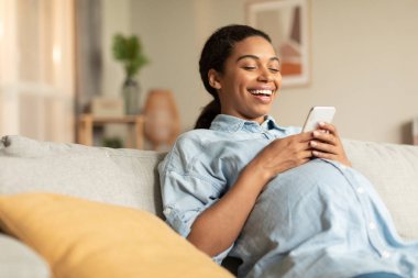 Joyful black pregnant woman using smartphone while relaxing on couch at home, happy lady expecting baby watching funny videos and laughing, copy space
