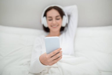 Modern mobile phone in young woman hand, happy relaxed lady enjoying lazy weekend at home, lying in bed, using wireless headphones and cell phone, watching movie online, bedroom interior, copy space