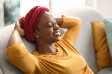 Closeup of happy smiling millennial african american woman wearing red headscarf chilling at home, sit on couch with closed eyes with hands behind head, dreaming about prosperous future, free space