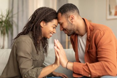 Romantic african american spouses holding their hands, touching foreheads, being intimate and affectionate, sitting on sofa at home. Loving black husband and wife bonding