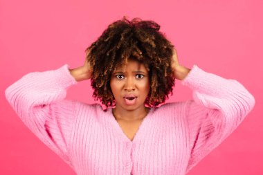 Shocked confused pretty millennial african american curly lady in casual with open mouth presses her hands to head isolated on pink background, studio. Human emotions, amazing bad news, ad and offer