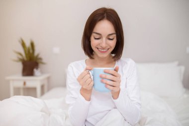 Happy beautiful brunette young woman in white pajamas sitting in bed, holding blue mug and smiling, cheerful lady drinking coffee in home bedroom after waking up in the morning, copy space