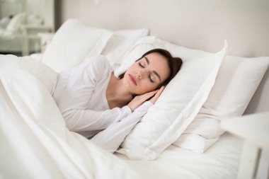 Beautiful young brunette woman sleeping with hands under her head on soft comfy pillows, peaceful female lying in bed with closed eyes, candid photo. Resting, sleeping, relaxation at home concept