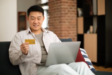 Happy mature asian man using credit card for online shopping, sitting on sofa with laptop computer, living room interior, copy space. Excited korean male paying for goods and services online