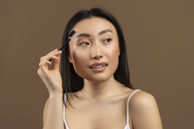 Beautiful young japanese lady brushing brows and applying eyebrow mascara, posing over brown studio background. Wellness and beauty concept